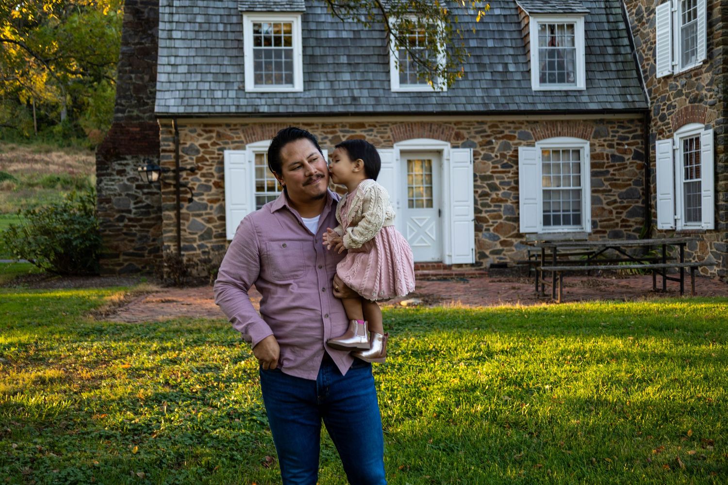 Man holding toddler girl in pink dress outside historic stone house on green lawn.