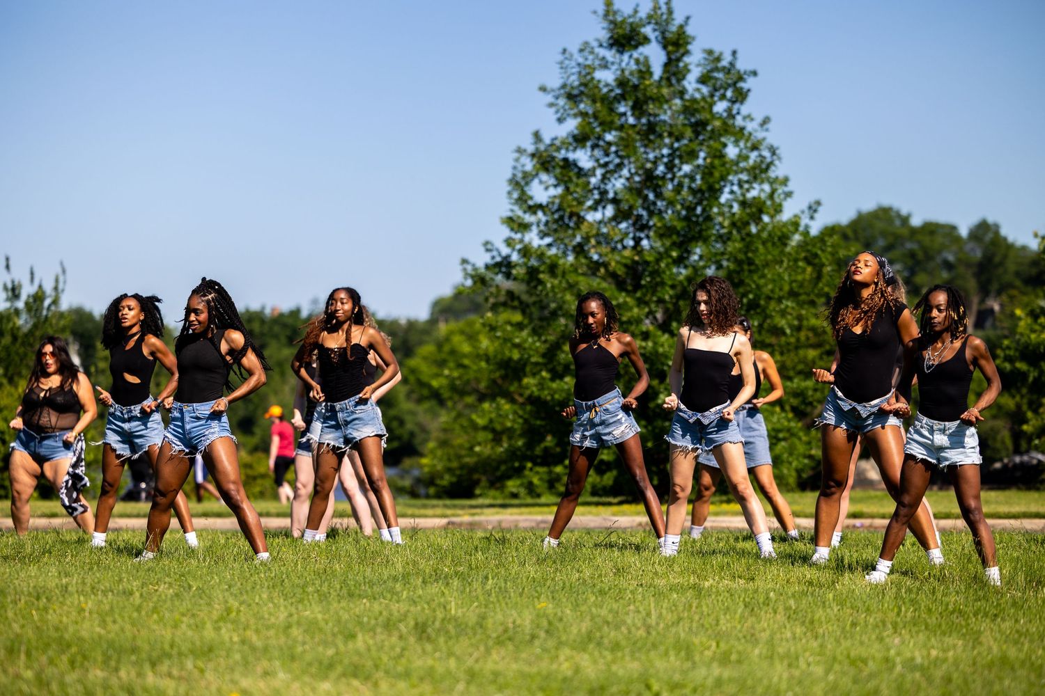 Group of young women in black tops and denim shorts performing a synchronized dance routine on a green field outdoors.
