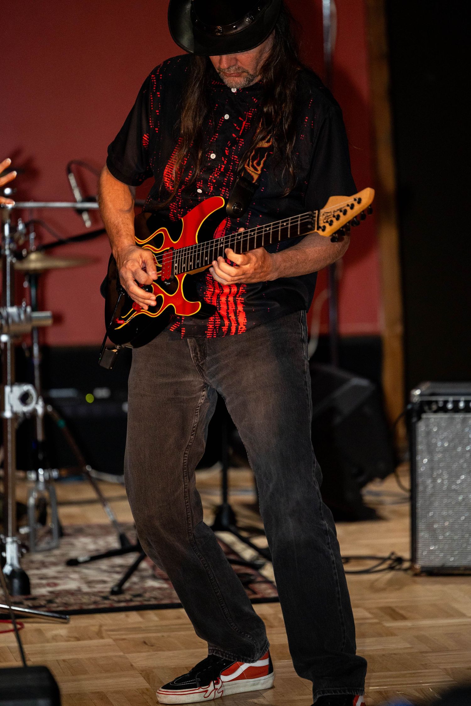 Male guitarist with long hair plays red flame electric guitar on stage wearing black hat and Vans shoes.