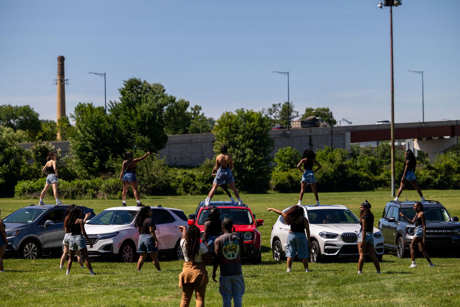 Cheerleaders performing stunts on cars and grass field outdoors on a sunny day.