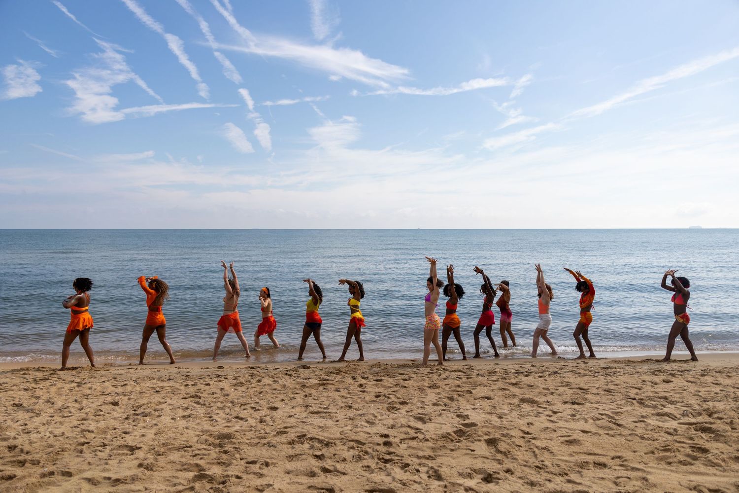 A line of diverse dancers performing choreography on a sandy beach with calm ocean waves and blue sky behind them.