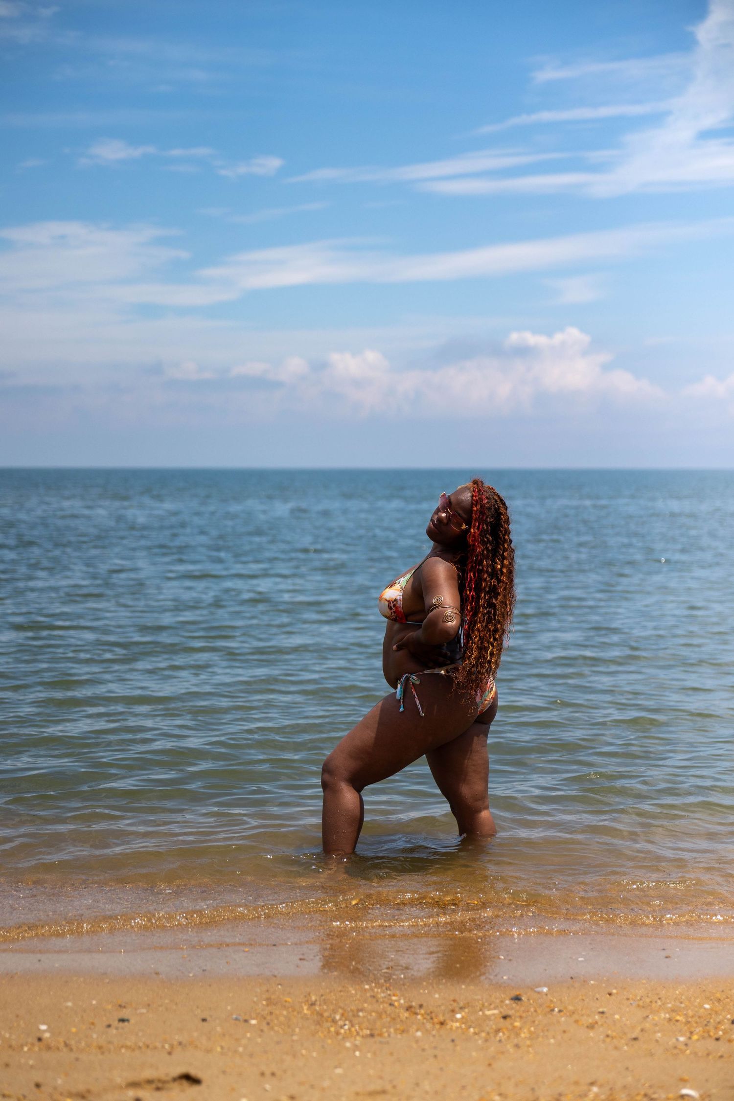 Woman with long braids in bikini standing in shallow ocean water on a sunny beach day.