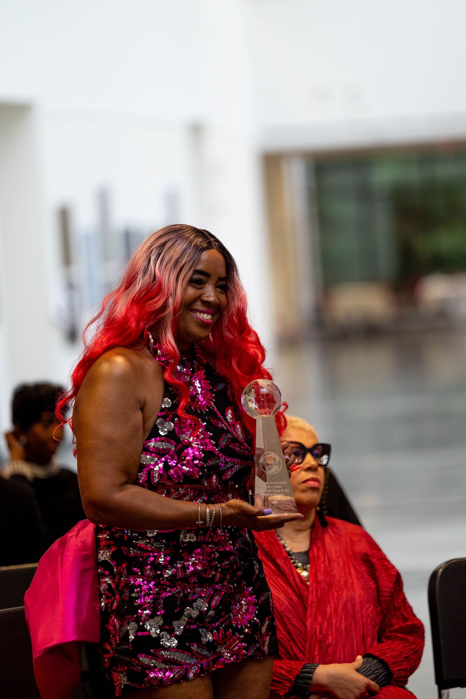 Woman in pink sequin dress holding crystal award trophy, smiling at an indoor awards ceremony event.