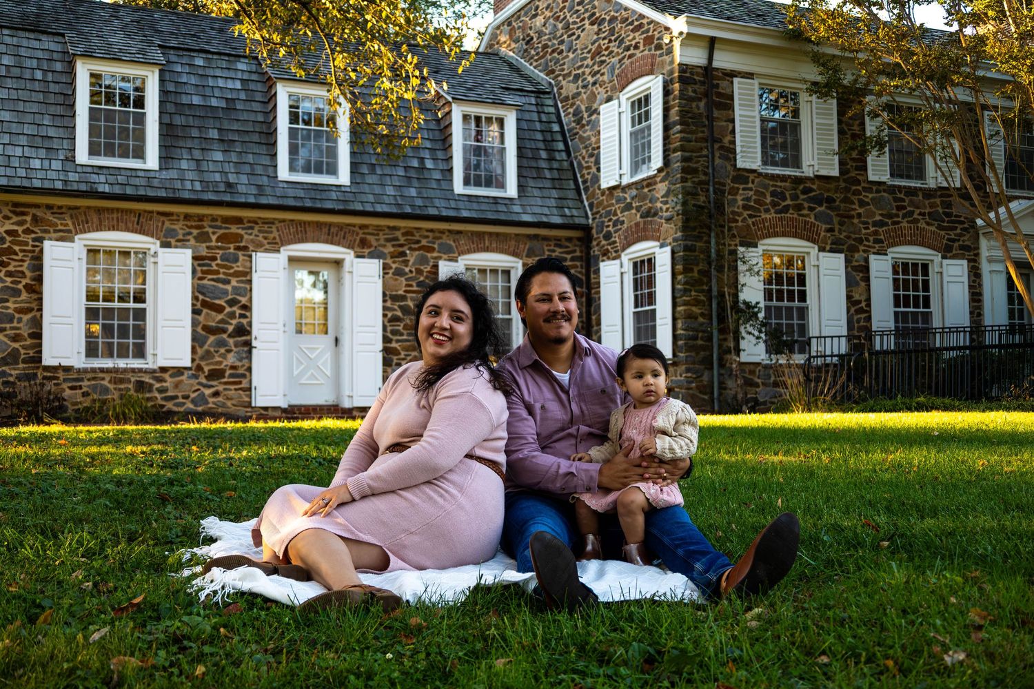 Happy family of three sitting on a blanket on green grass in front of a charming brick colonial home.