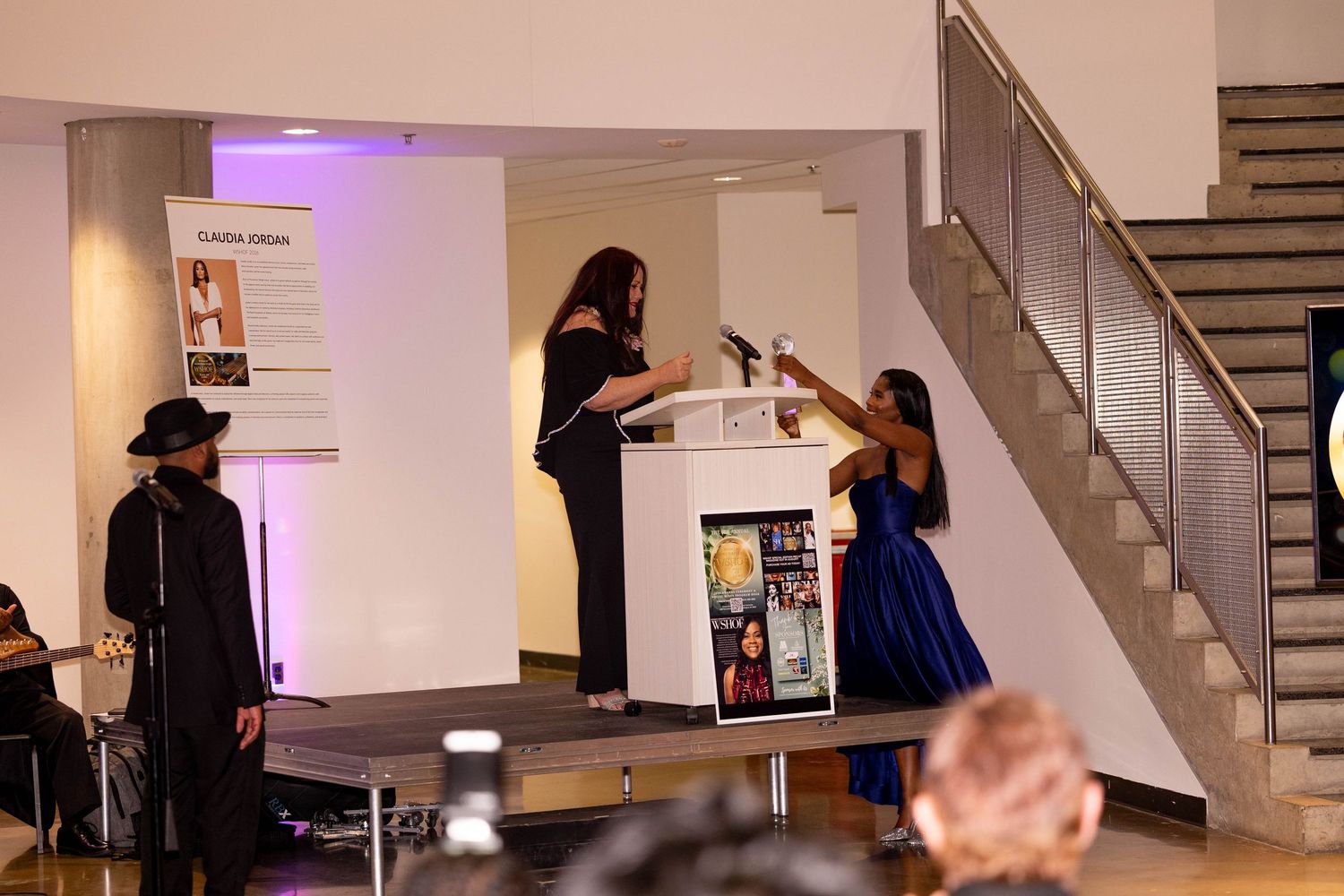 Two women at podium during Claudia Jordan event with performer on stage.