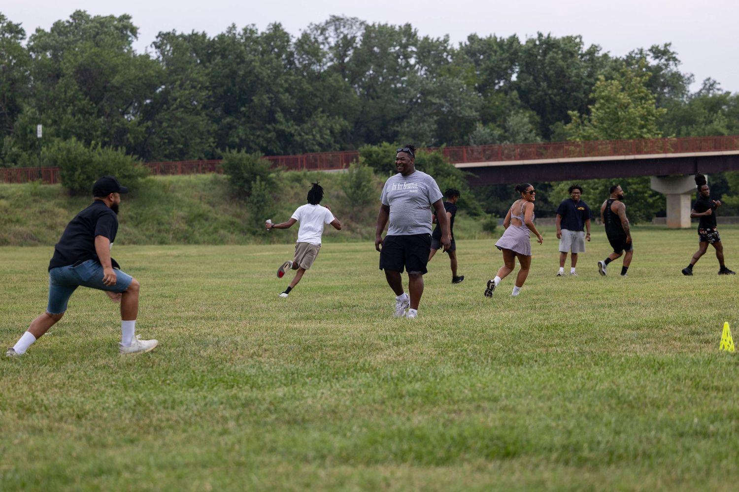 Group of people playing flag football on a green grass field outdoors near trees and a bridge.