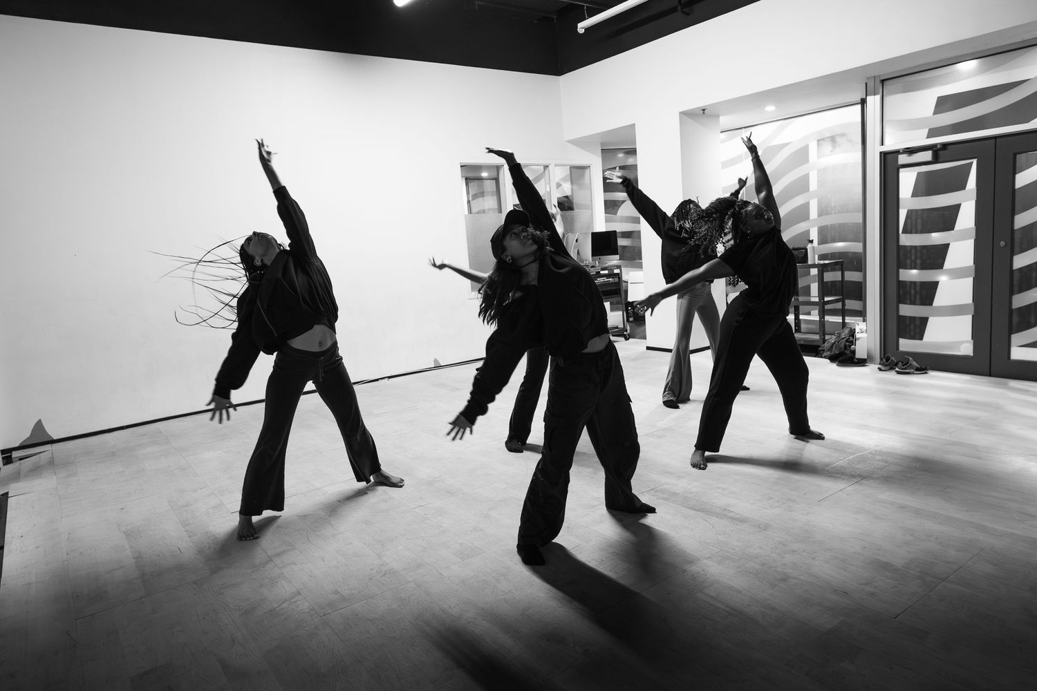 Black and white photo of four dancers in dark outfits performing contemporary dance moves in a studio.