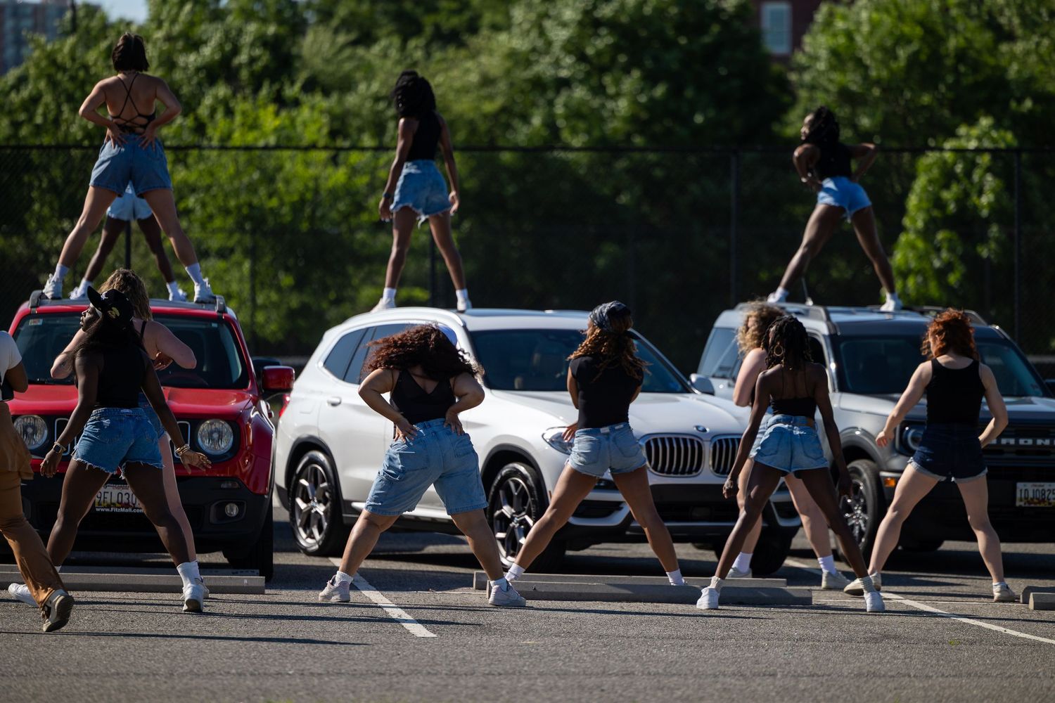 A group of young women in denim shorts perform a synchronized dance routine in a parking lot surrounded by cars.