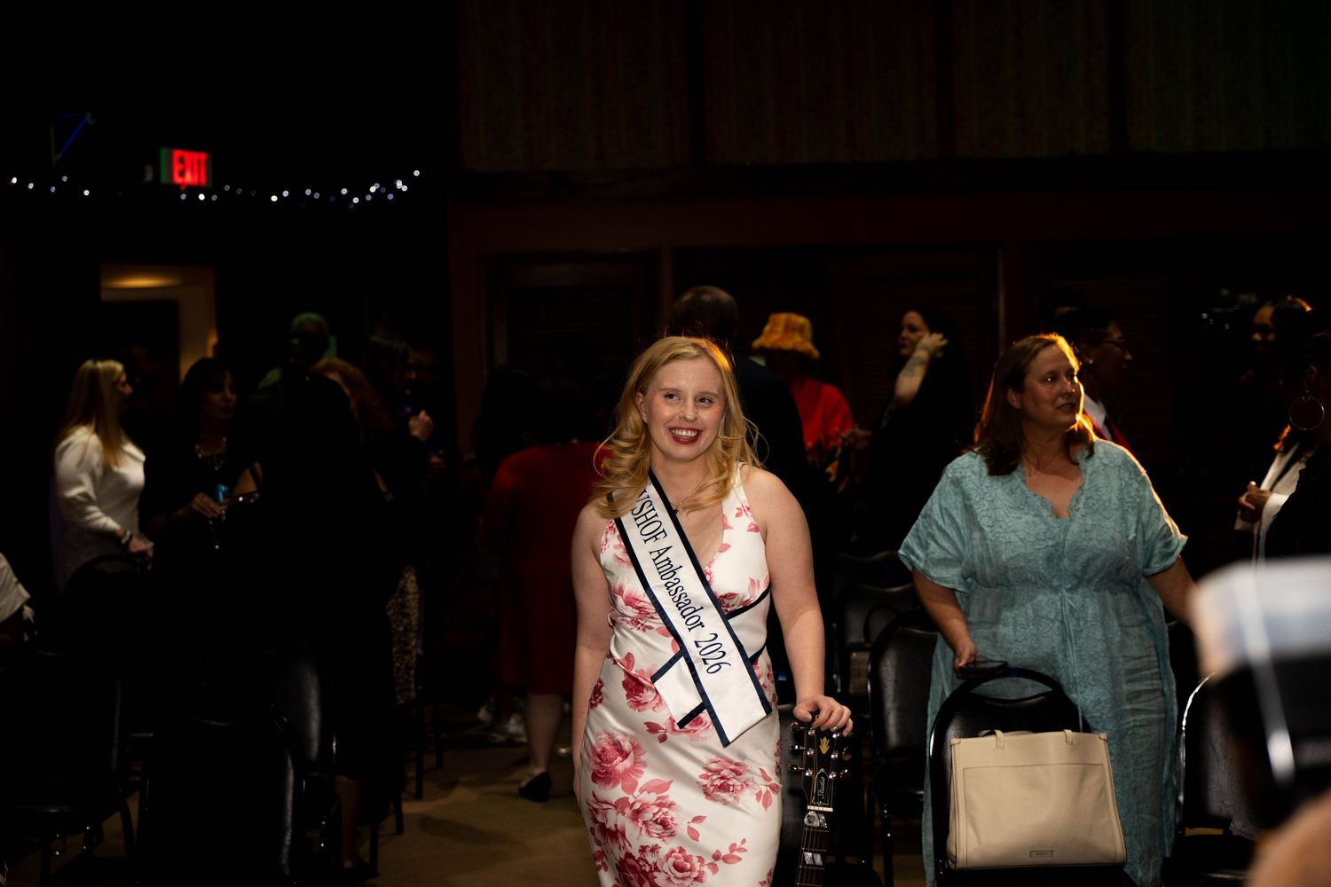 Young woman in floral dress wearing pageant sash walks runway at formal event with crowd in background.