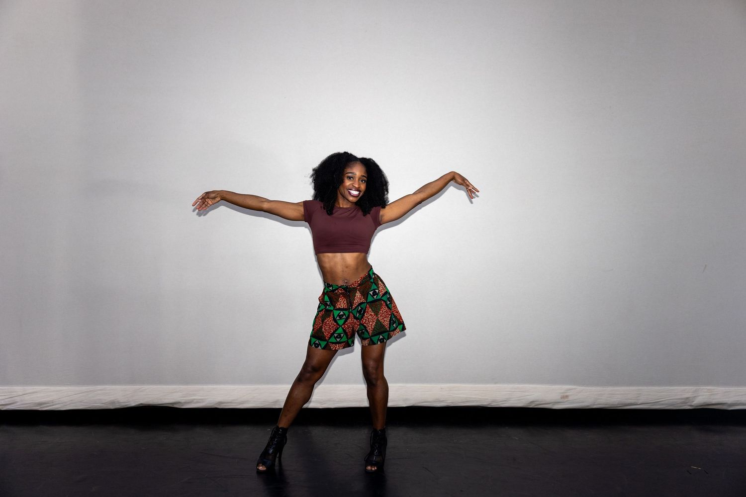 A woman in a red crop top and colorful printed skirt dances joyfully with arms outstretched against a gray background.