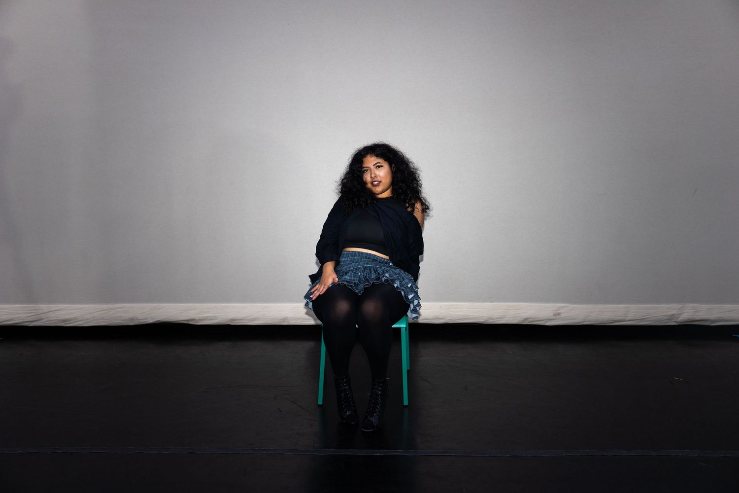 A young woman with curly hair sits smiling on a green stool against a gray wall in a black sweater and patterned skirt.