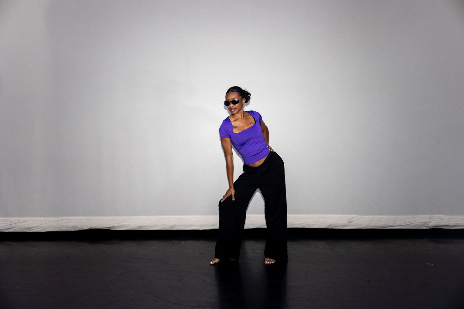 A woman in a purple top and black pants performs an expressive dance move in a bright studio space.