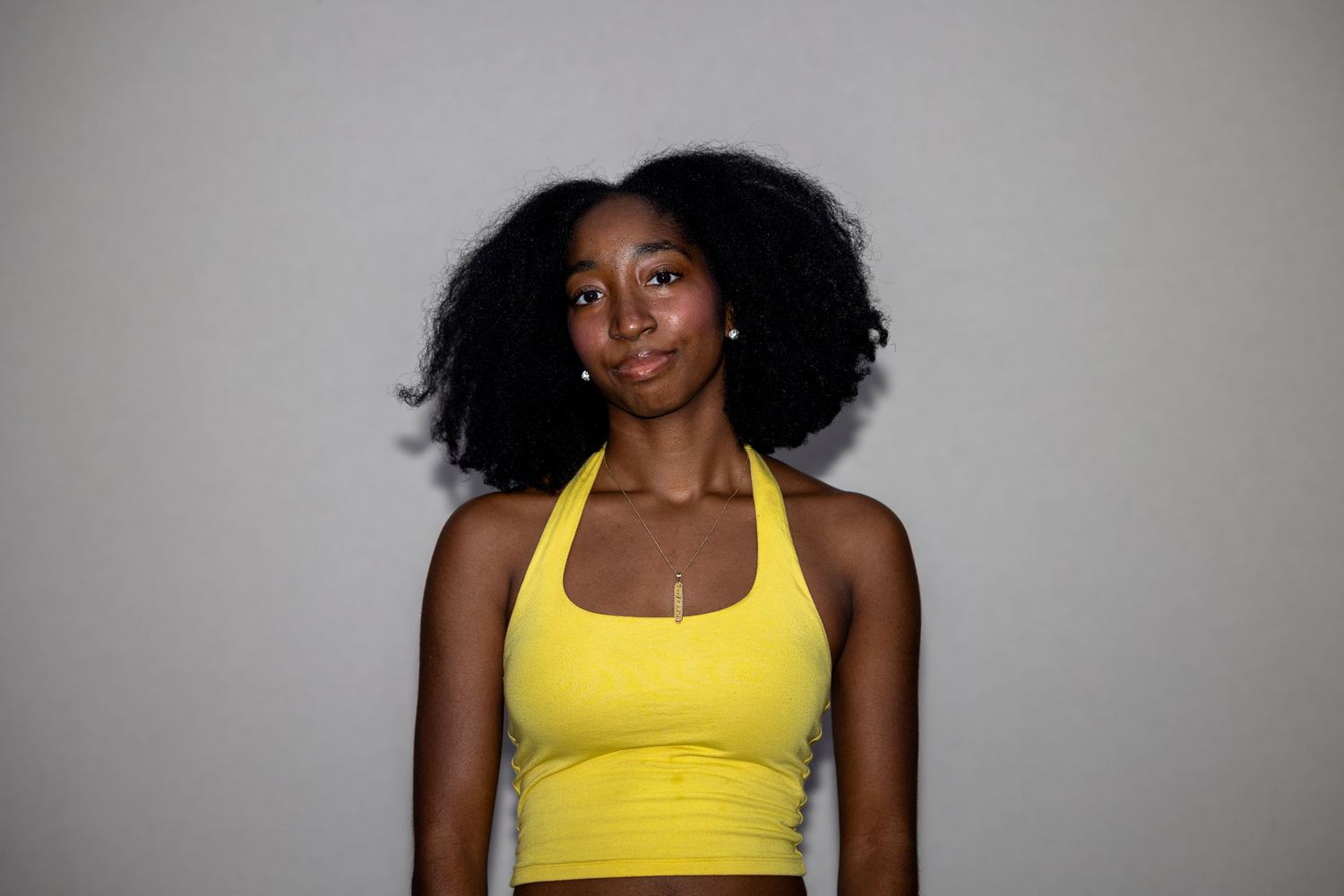 Young woman with natural curly hair wearing a yellow halter crop top posing against a light gray background.
