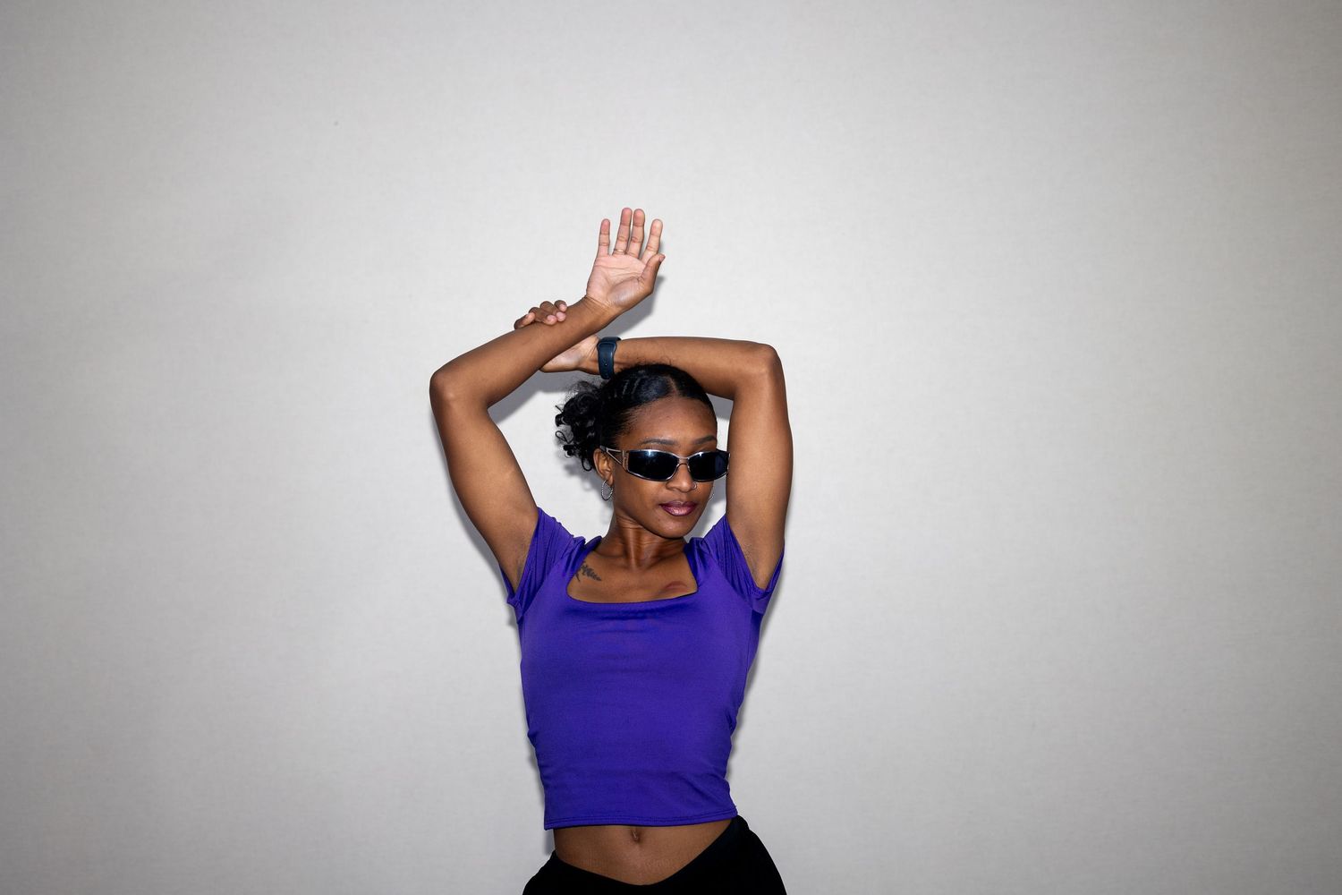 Woman in purple crop top and sunglasses dances with arms raised against white background.