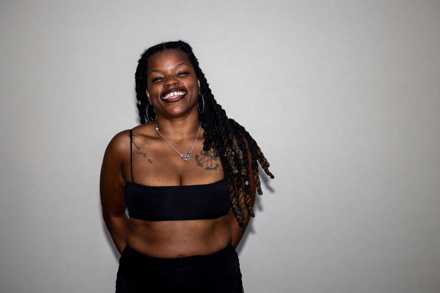 Laughing woman with long braids in black crop top and pants posing with crossed arms against white background.