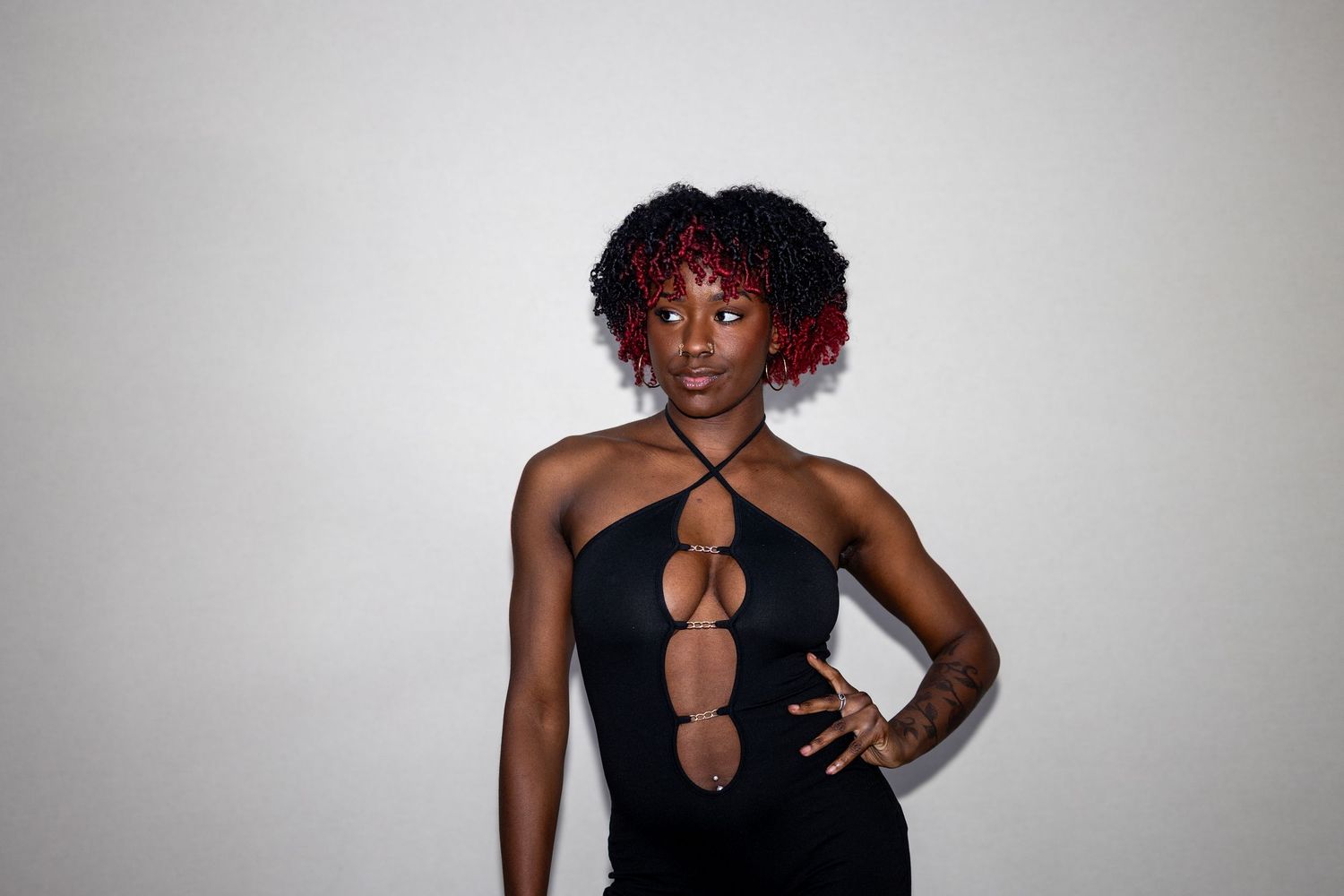 Woman in black cutout dress with red-tipped natural hair poses confidently against white background.