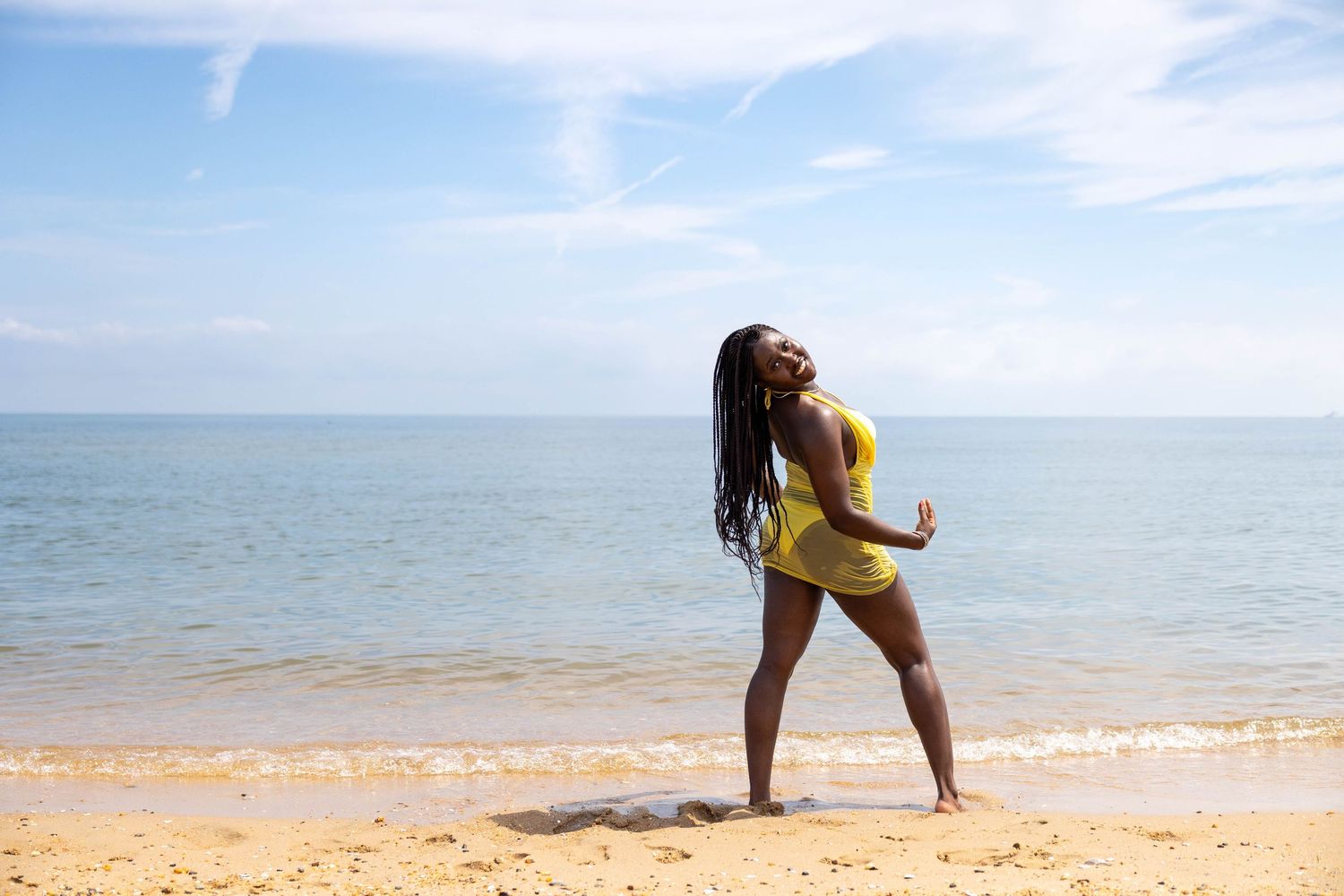 A young Black woman in a yellow dress poses joyfully on a sunny sandy beach with calm blue ocean waters behind her.