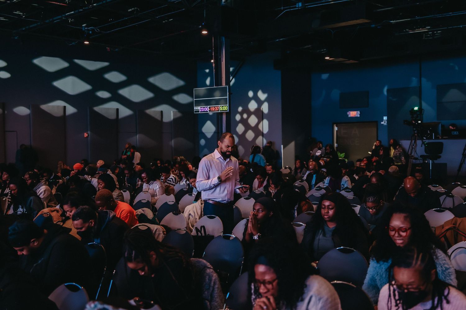 A crowded tech conference or meetup event with attendees seated in rows watching a presentation in a dark room.