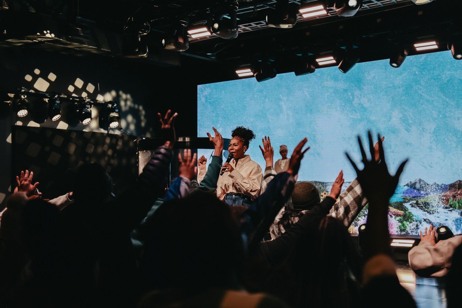 Concert crowd raises hands in silhouette against colorful stage lighting during live music performance.