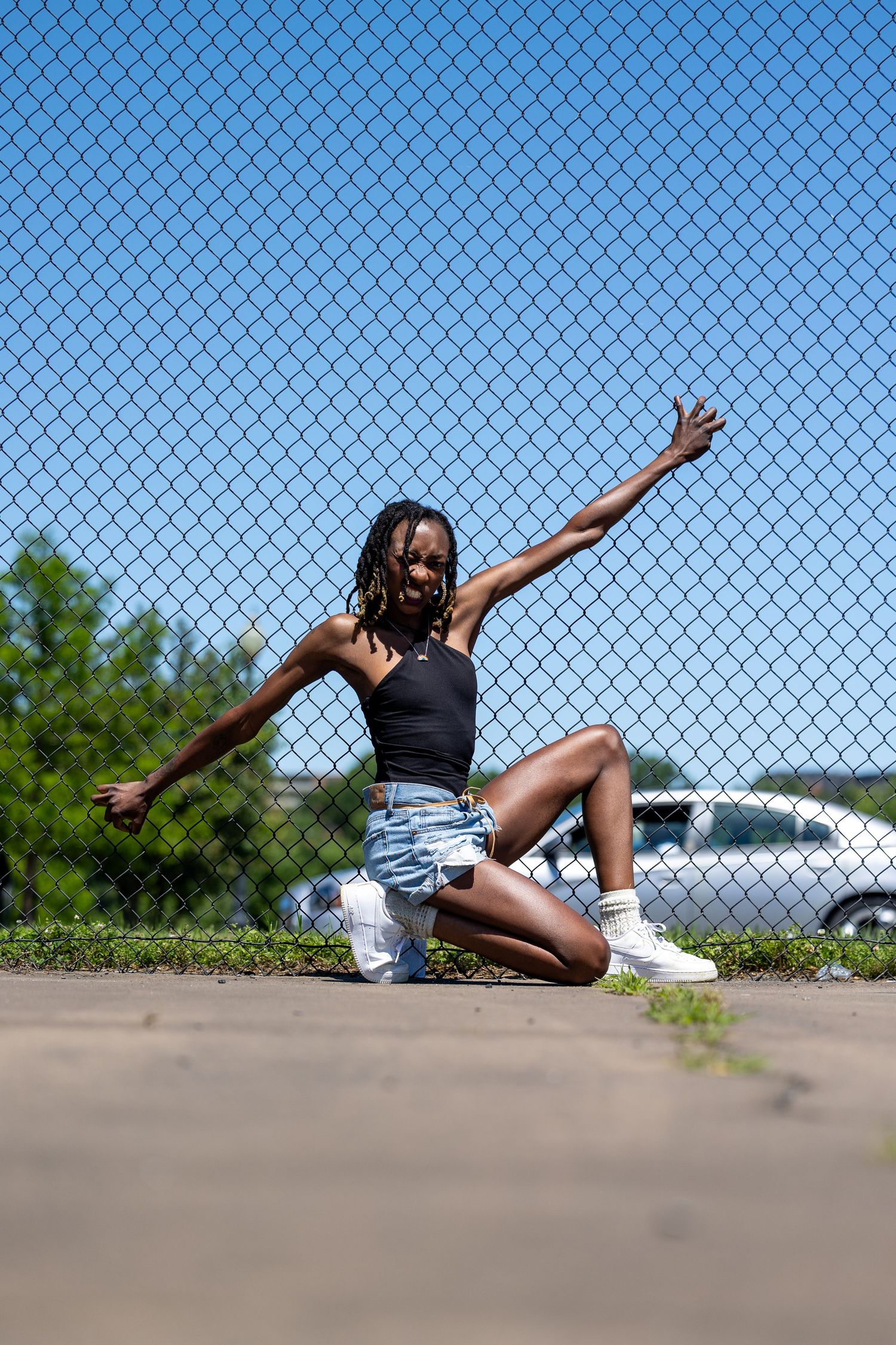 Young Black woman in black crop top and denim shorts poses dynamically on pavement before chain-link fence on sunny day.