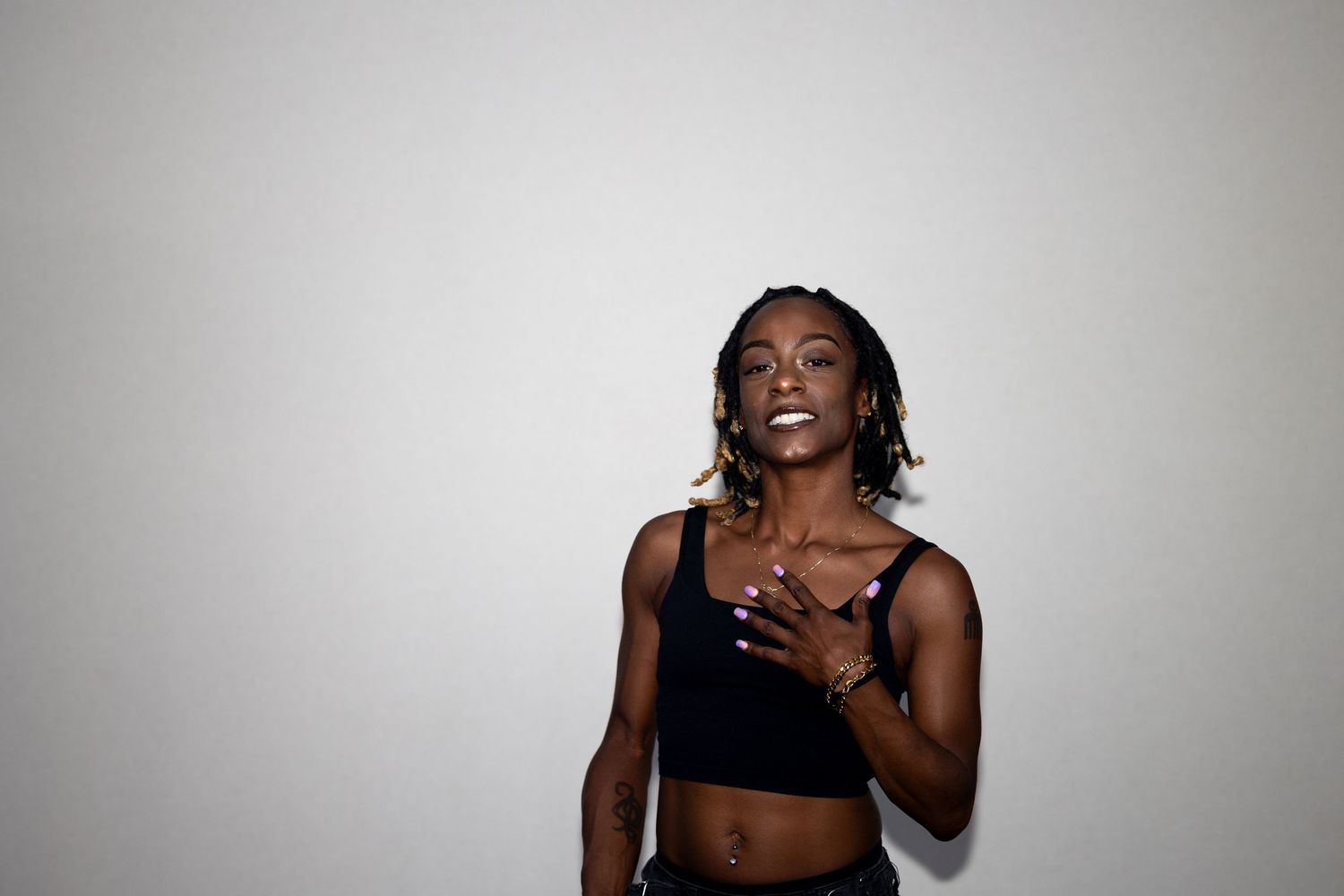 Laughing woman in black crop top with hand on chest wearing gold jewelry against white background.