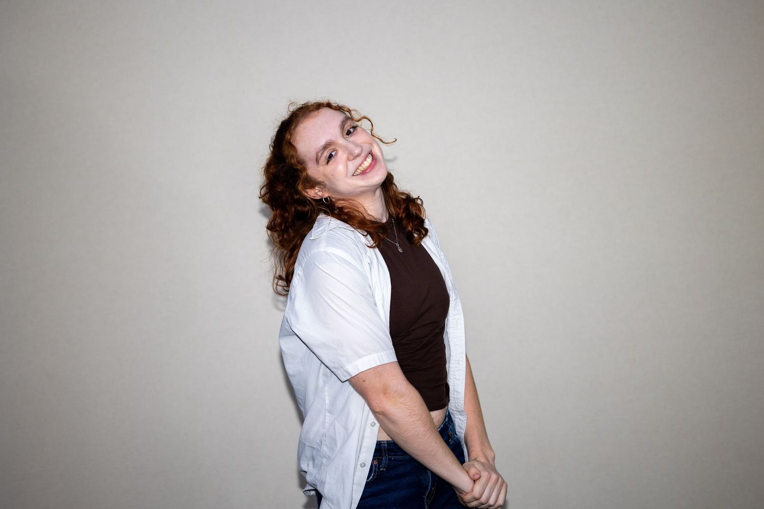 Curly-haired young woman in white jacket and dark top smiles over shoulder against gray background.