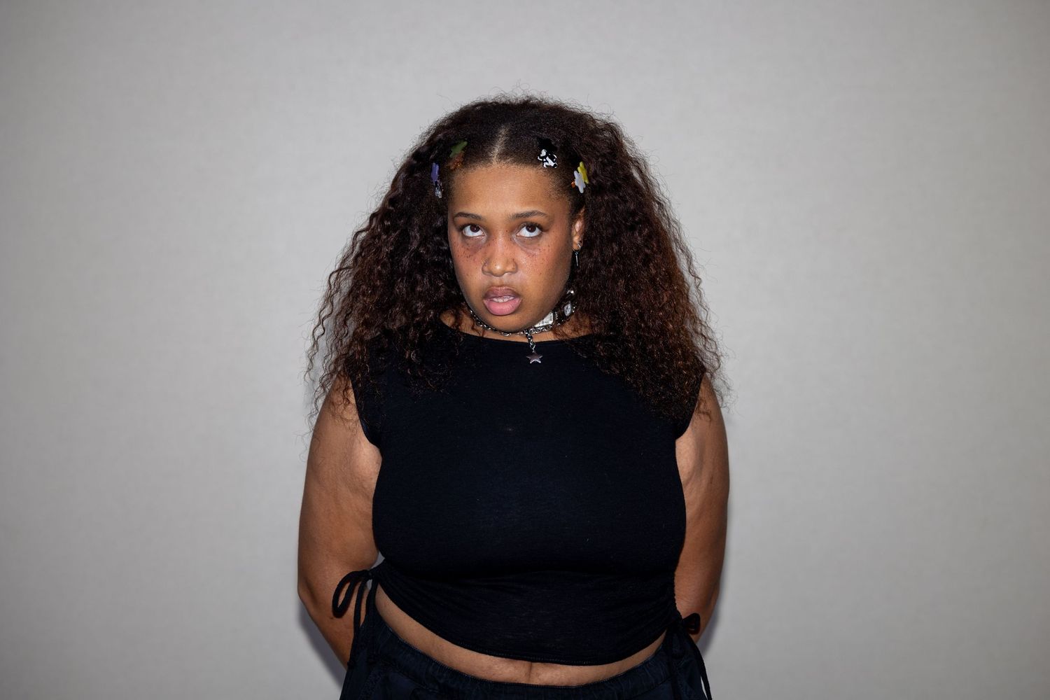 Young woman in black crop top with curly hair poses with arms crossed.