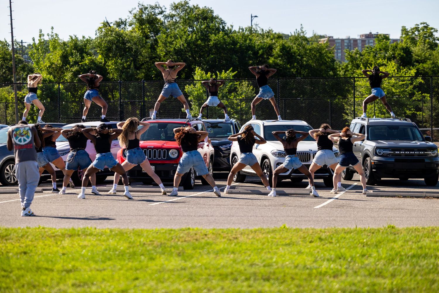 Cheerleaders performing stunts in a parking lot with cars and green trees in the background on a sunny day.