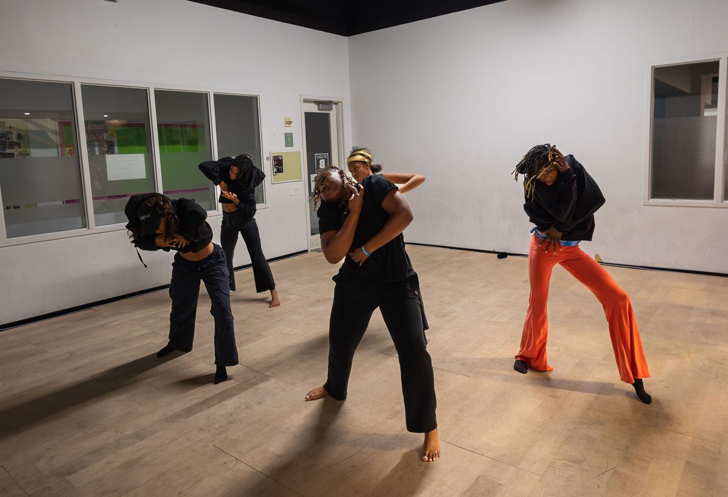 Four dancers perform expressive choreography in a modern studio with wooden floors and white walls.