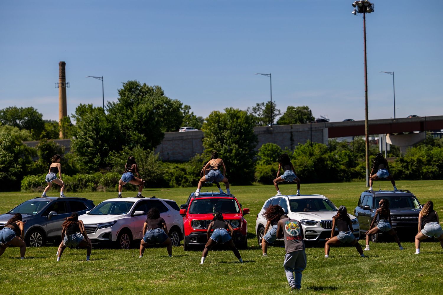 Performers dancing on and around colorful cars on a green field with trees and industrial buildings in the background.