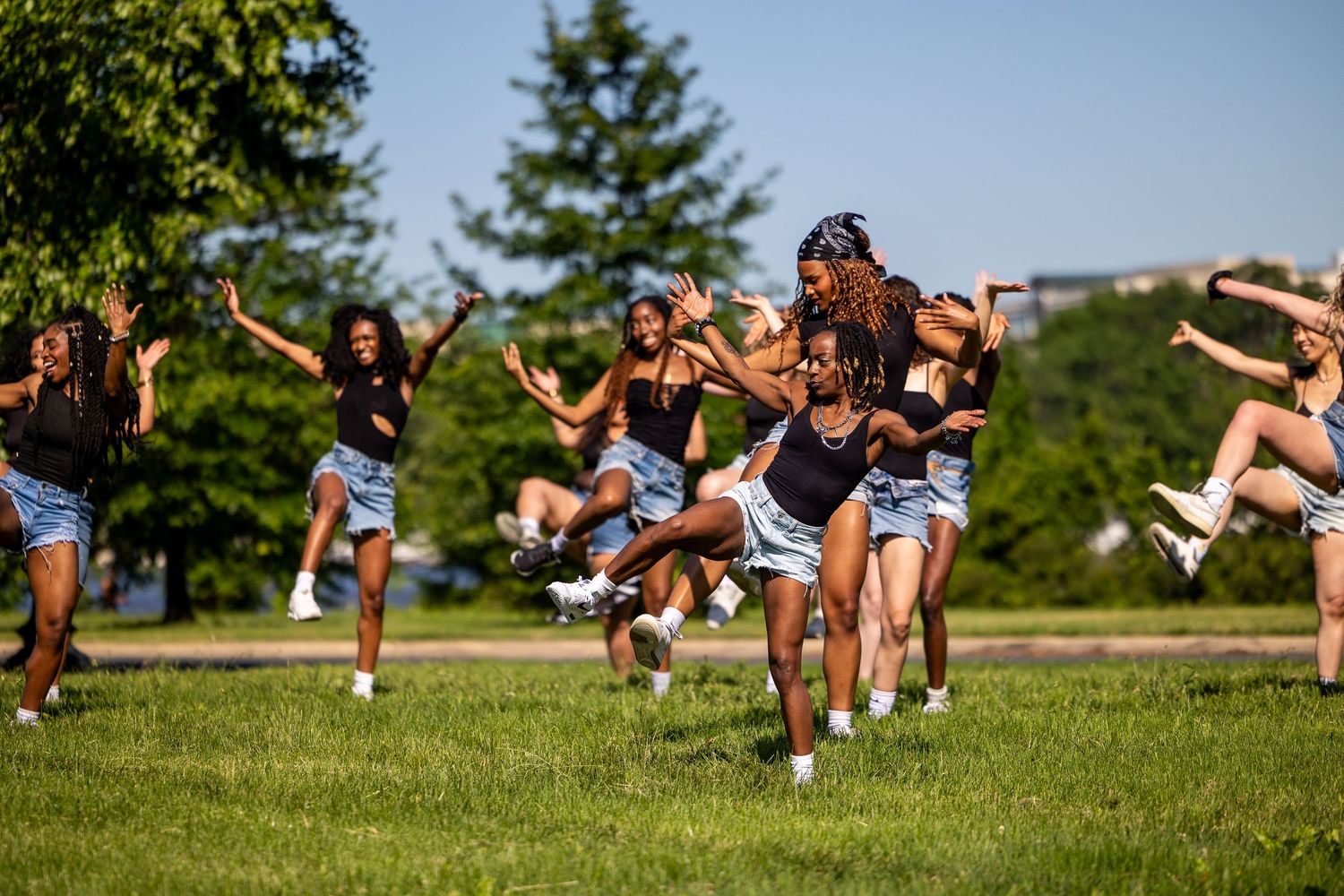 A group of young women dancing energetically outdoors on green grass with trees in the background on a sunny day.