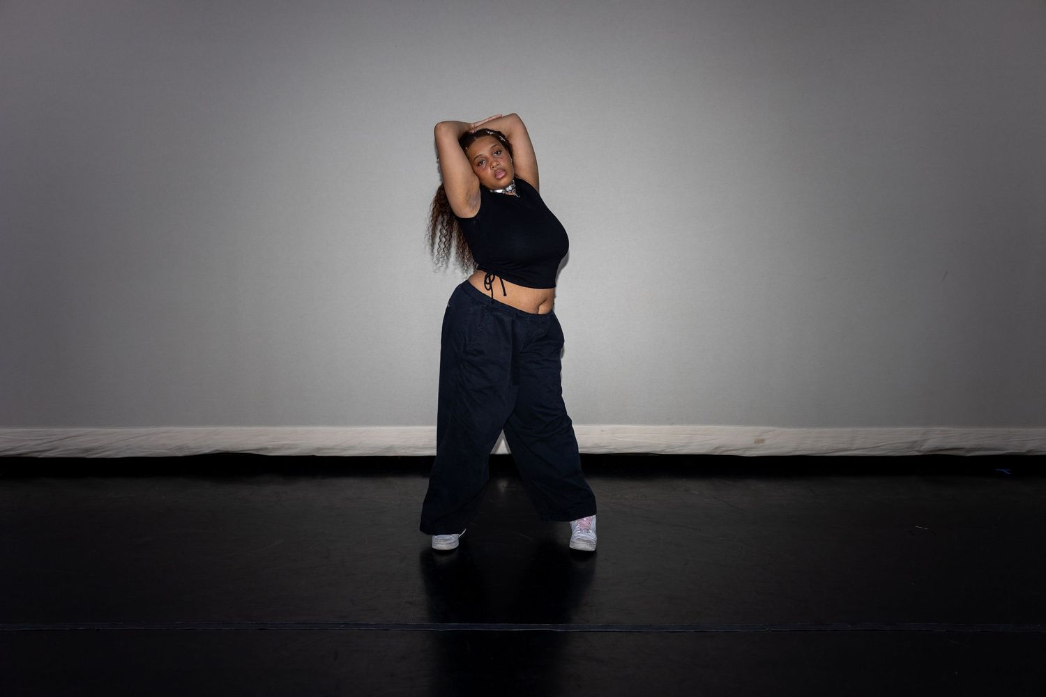 Woman in black crop top and wide-leg jeans dancing with arms raised overhead in a studio.