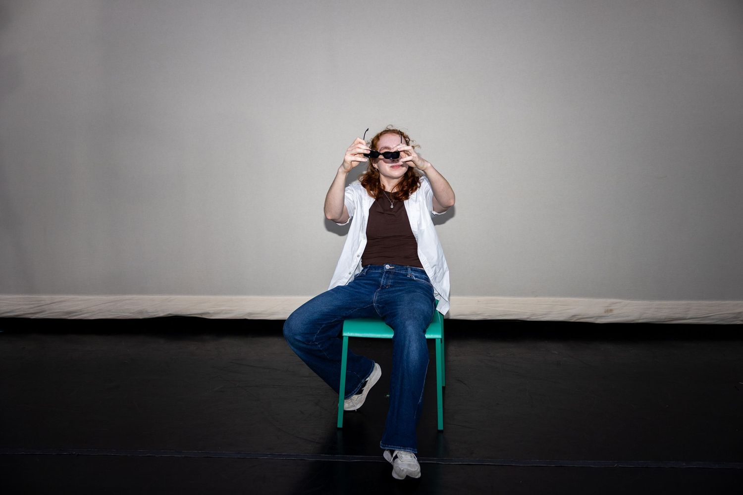Curly-haired performer sits on green chair adjusting sunglasses in white jacket and flare jeans.
