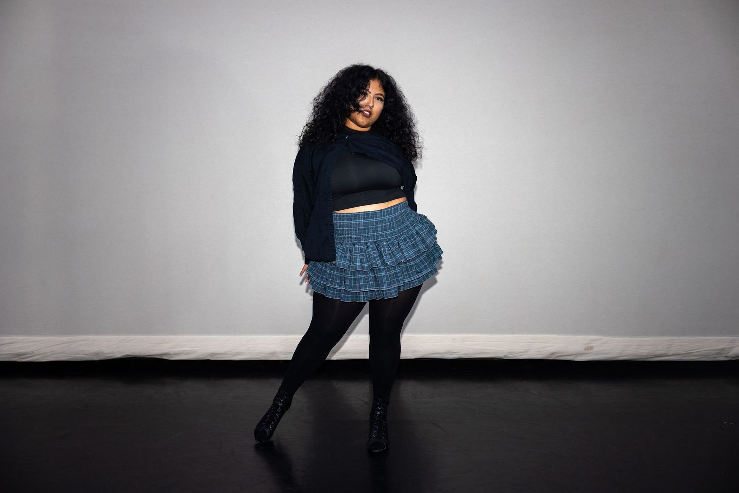 A curvy woman in black crop top and blue plaid mini skirt with tights poses confidently.