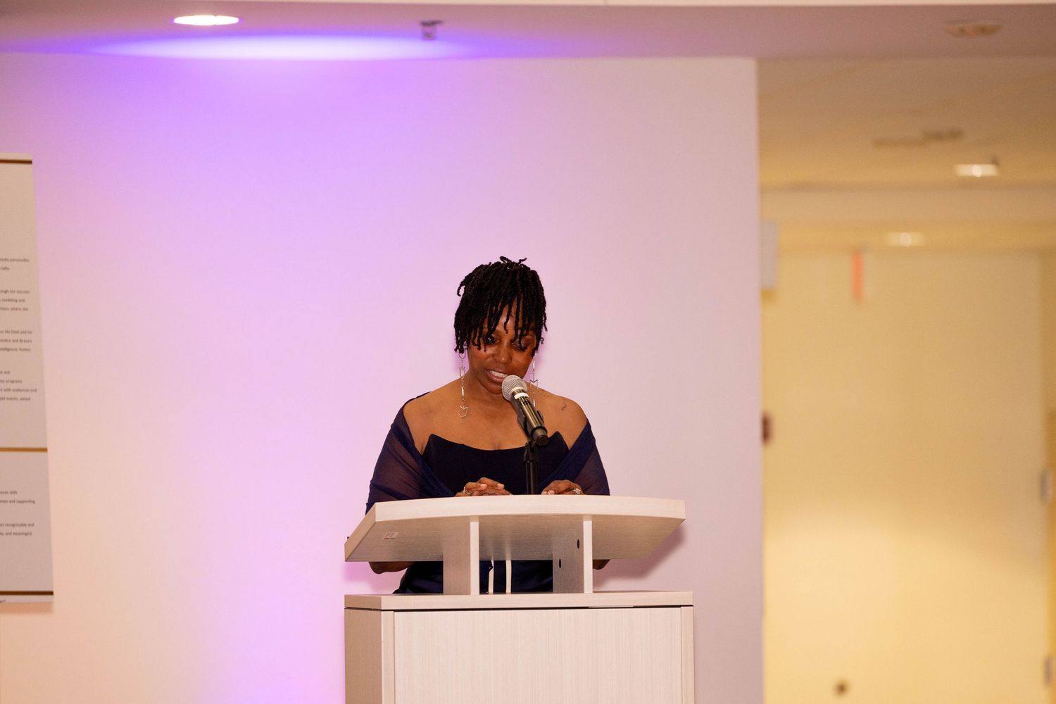 Woman speaking at a white podium during an indoor event with purple ambient lighting in the background.