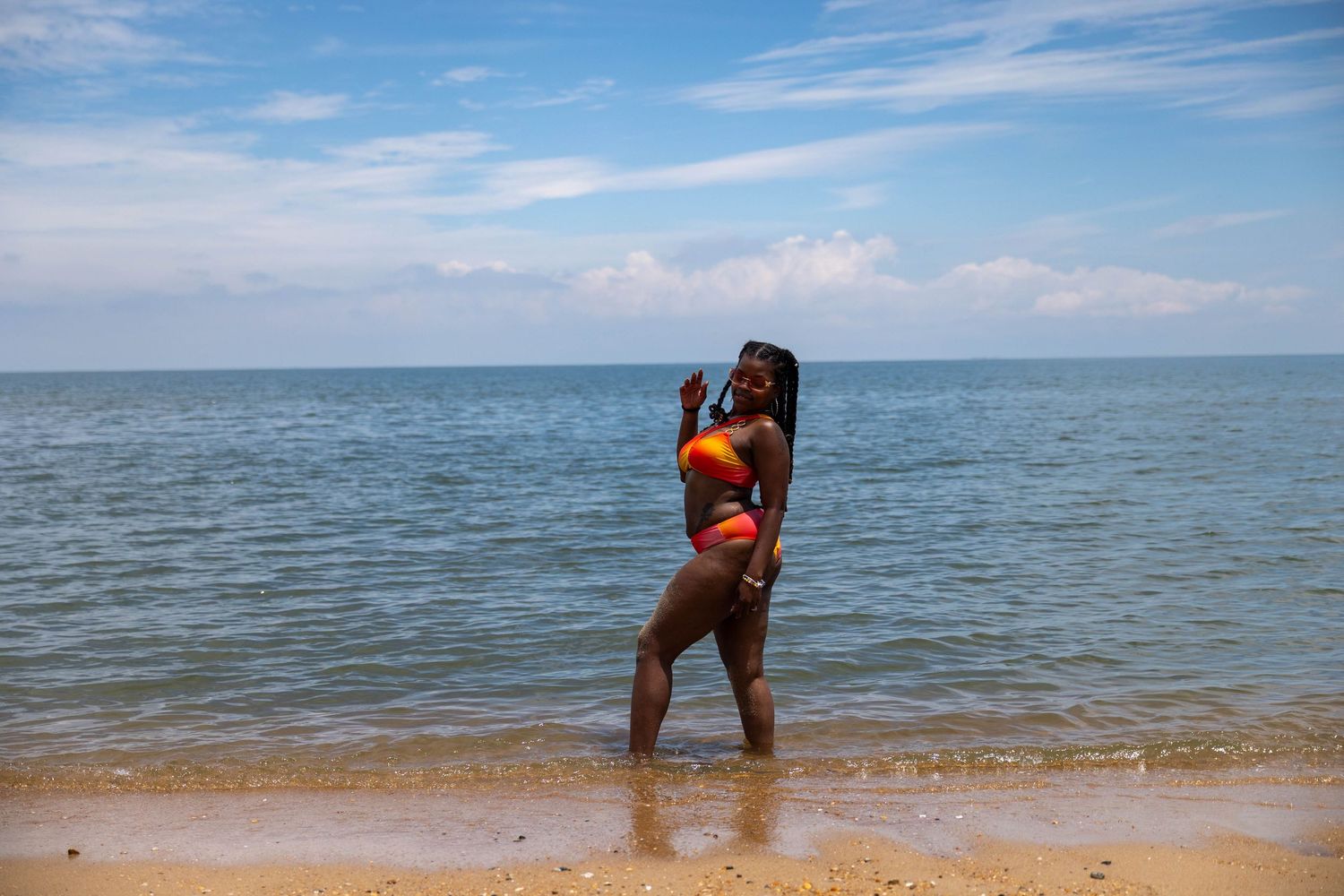 A Black woman in a vibrant red and yellow bikini poses at the ocean's edge on a sunny beach.