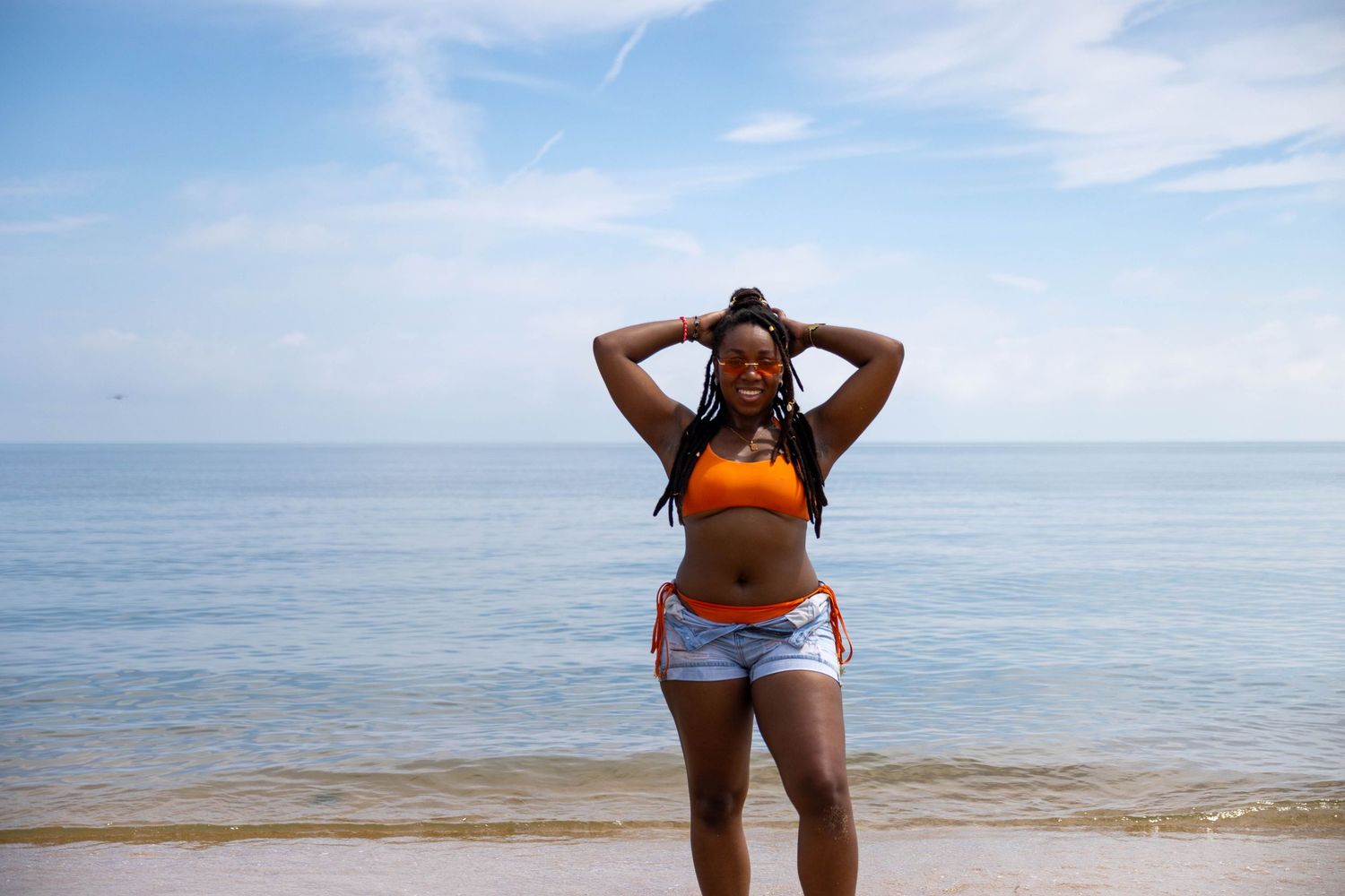 Woman in orange crop top and denim shorts posing confidently on a sunny beach with blue ocean water behind her.