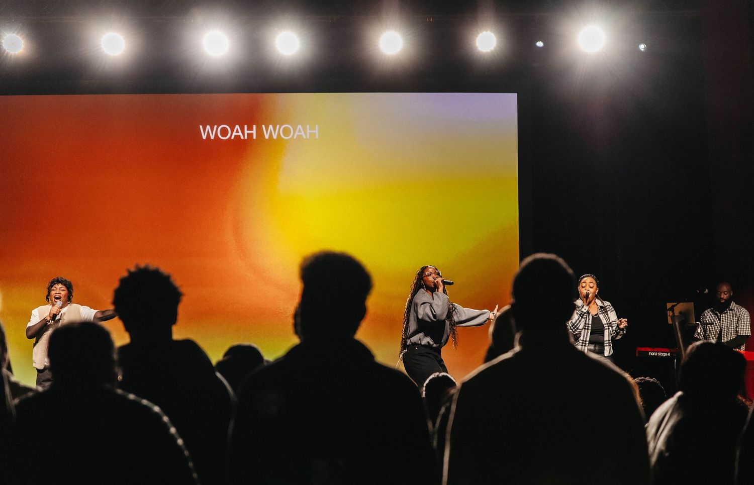 Concert stage with bright spotlights illuminating performers silhouetted against an orange and black backdrop.