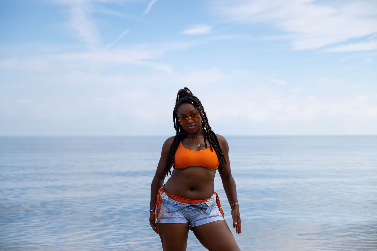 Young Black woman in orange crop top and denim shorts posing confidently on a beach with blue ocean backdrop.
