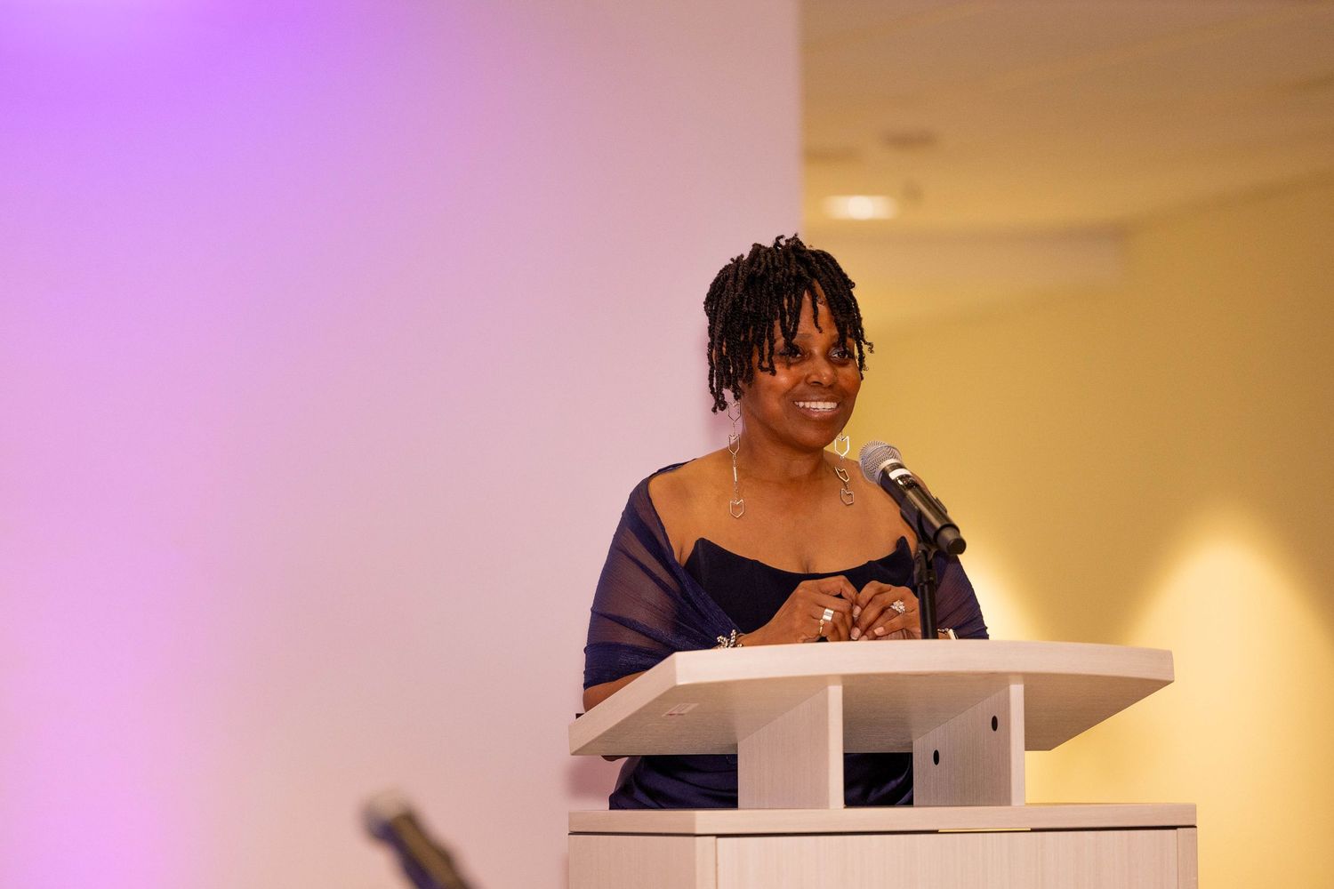 Woman with dreadlocks speaks at a white podium with microphone at an indoor event.