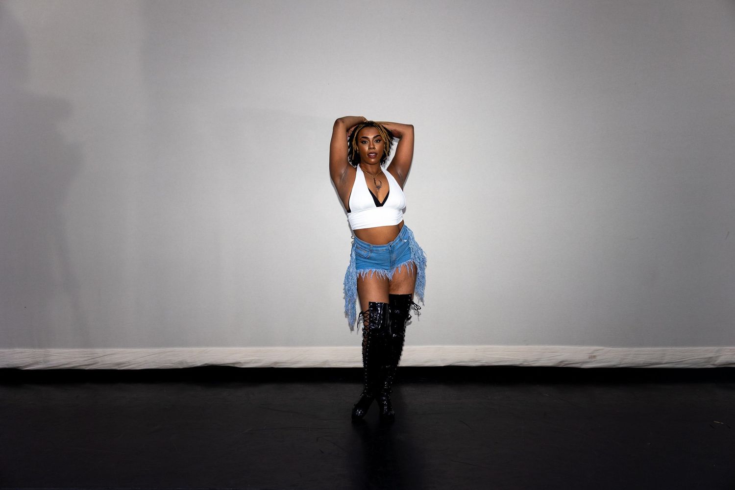 Woman in white crop top, denim fringe skirt, and thigh-high boots poses in a studio.
