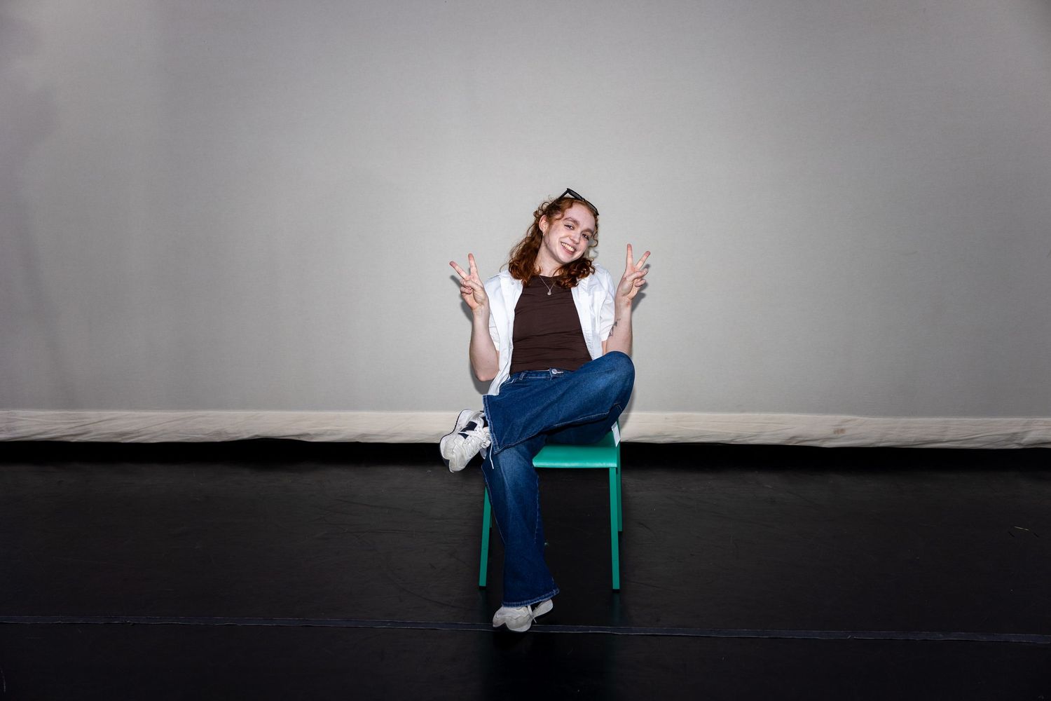 A cheerful young woman in jeans and brown top sits on a green chair making peace signs with both hands.