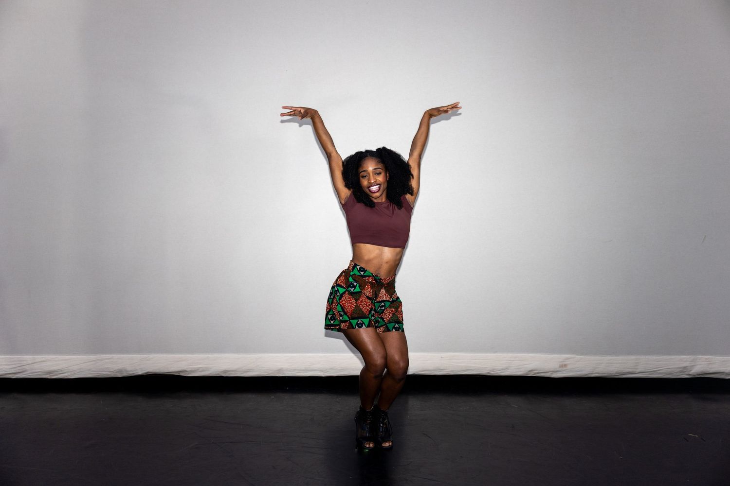 Joyful dancer in floral skirt and crop top raises arms with bright smile in studio setting.
