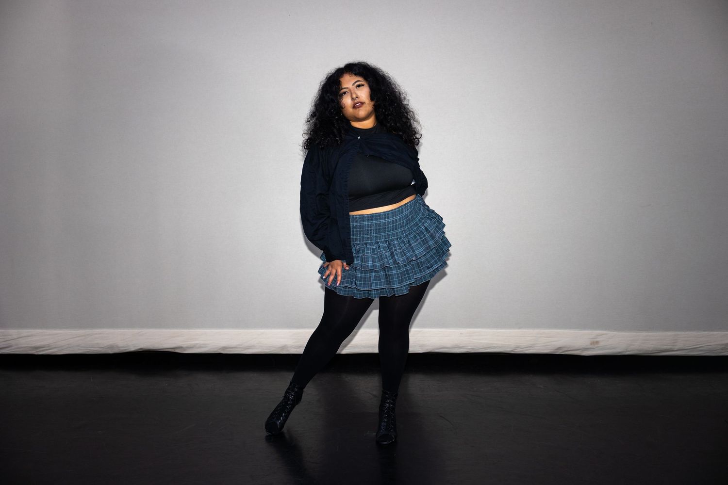 Woman in blue plaid mini skirt and black sweater posing in a dance studio.