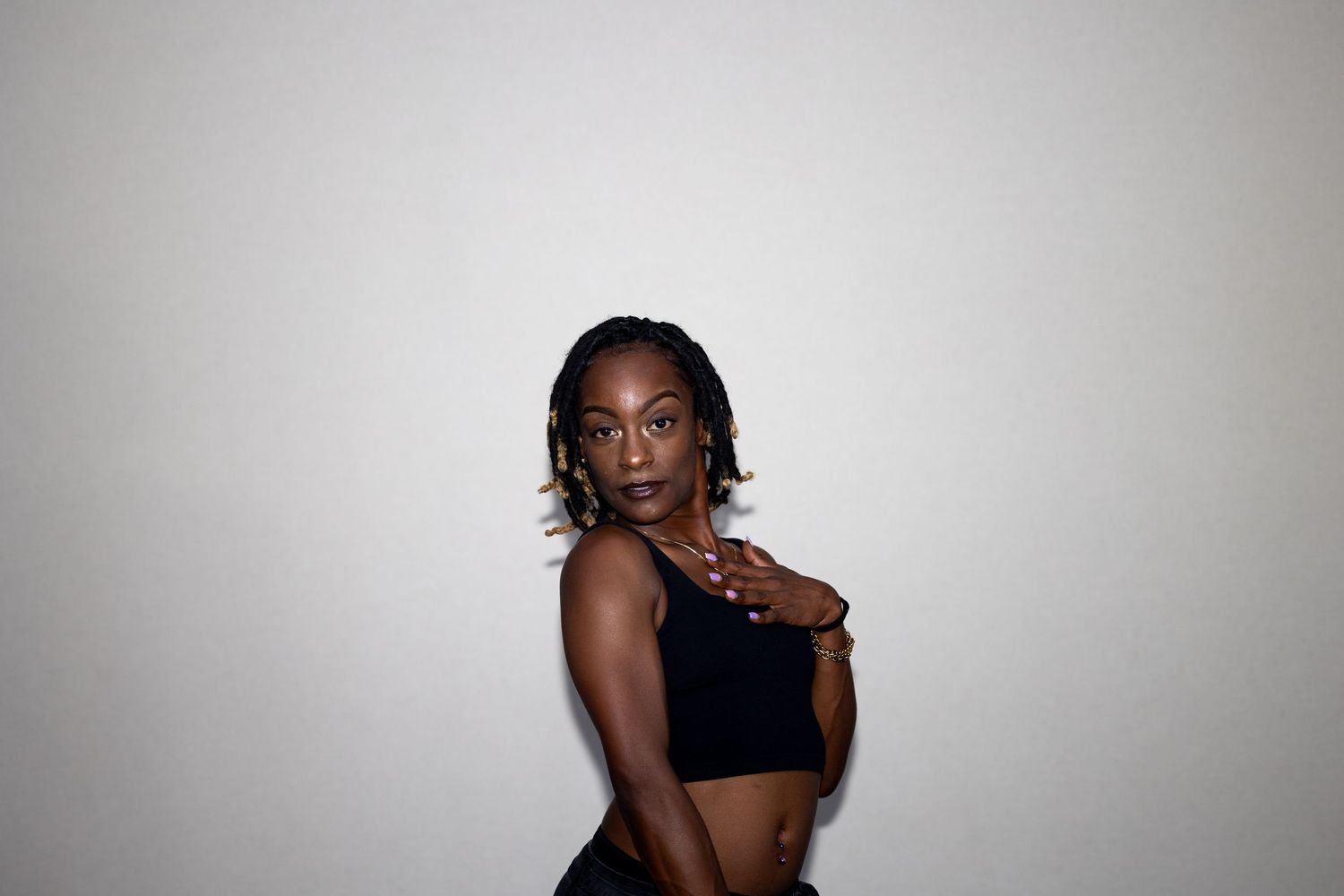 A young woman in a black crop top gazes over her shoulder with a serious expression against a gray wall.