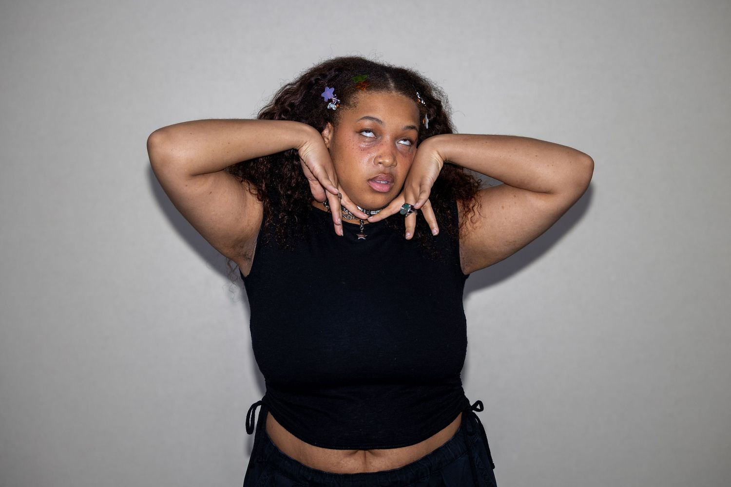 Young woman in black crop top posing with hands raised near face, purple hair clips, against gray background.