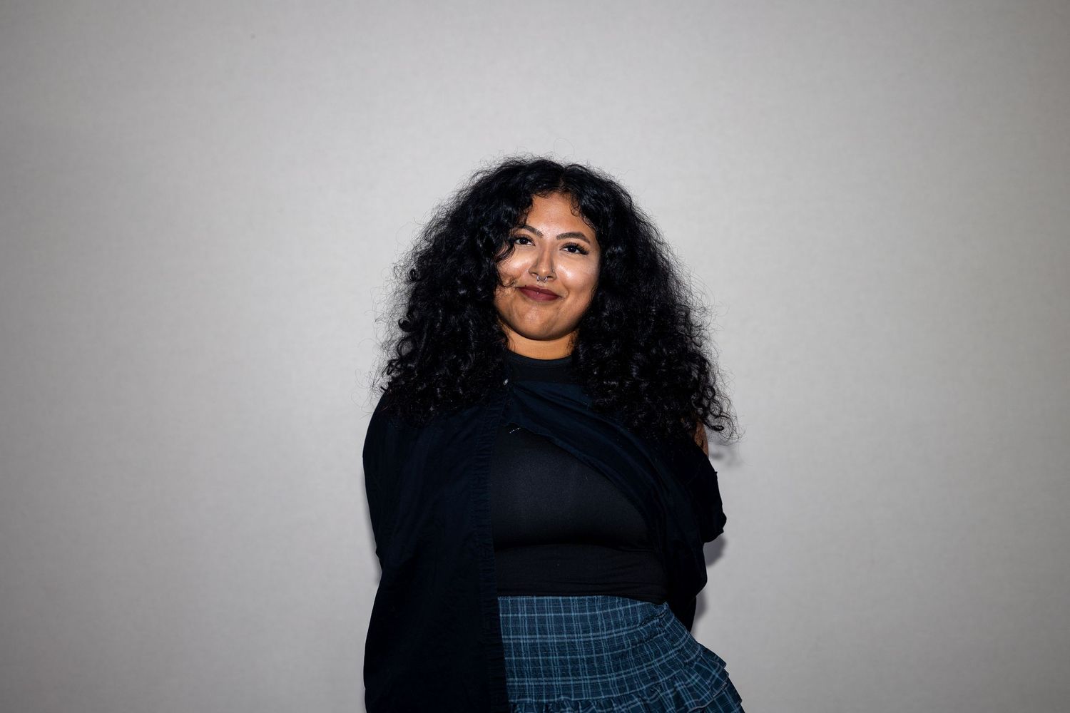 Woman with curly black hair wearing black turtleneck and plaid skirt smiling over shoulder against gray background.