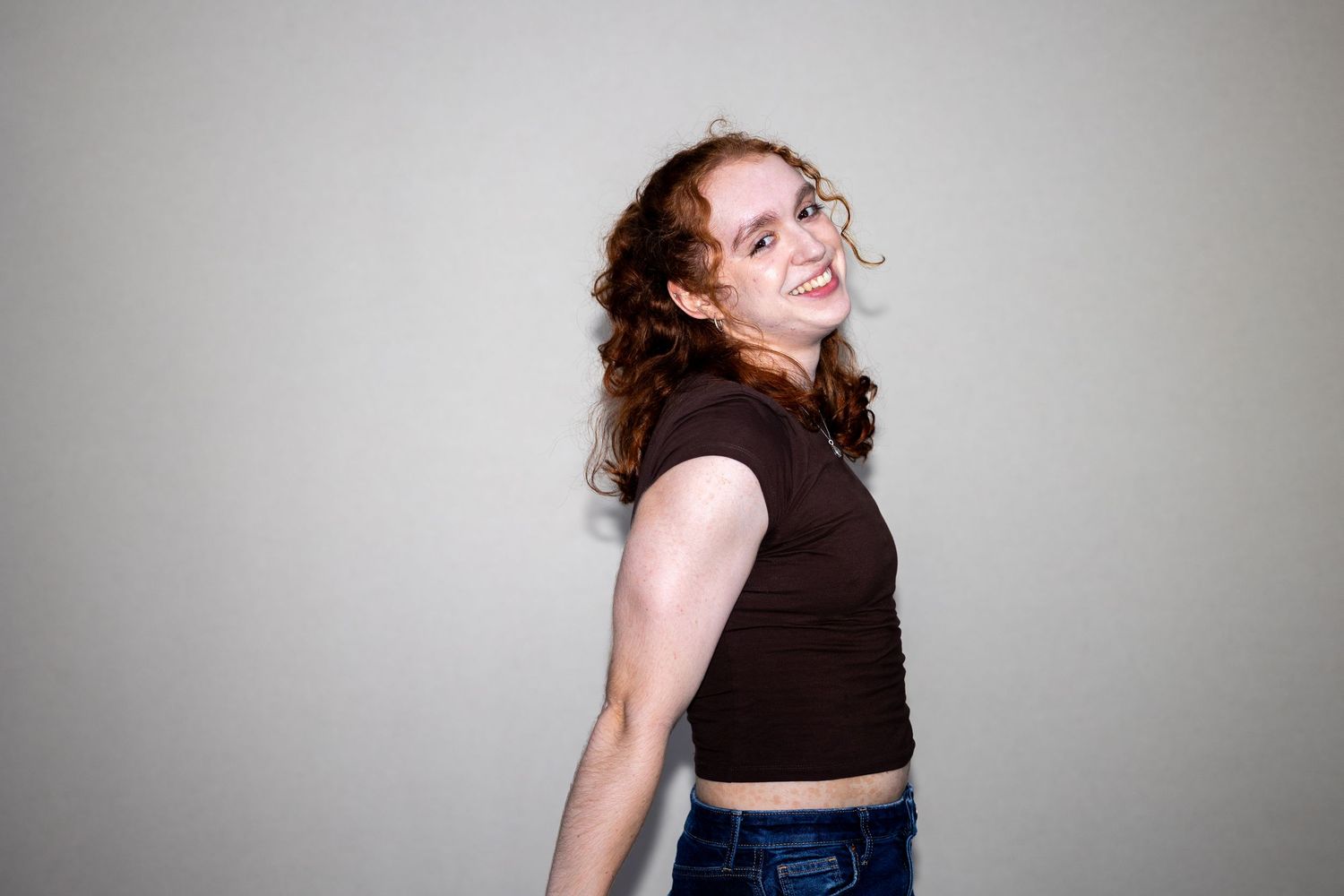 A woman with curly red hair smiles over her shoulder in a dark brown crop top and blue jeans.