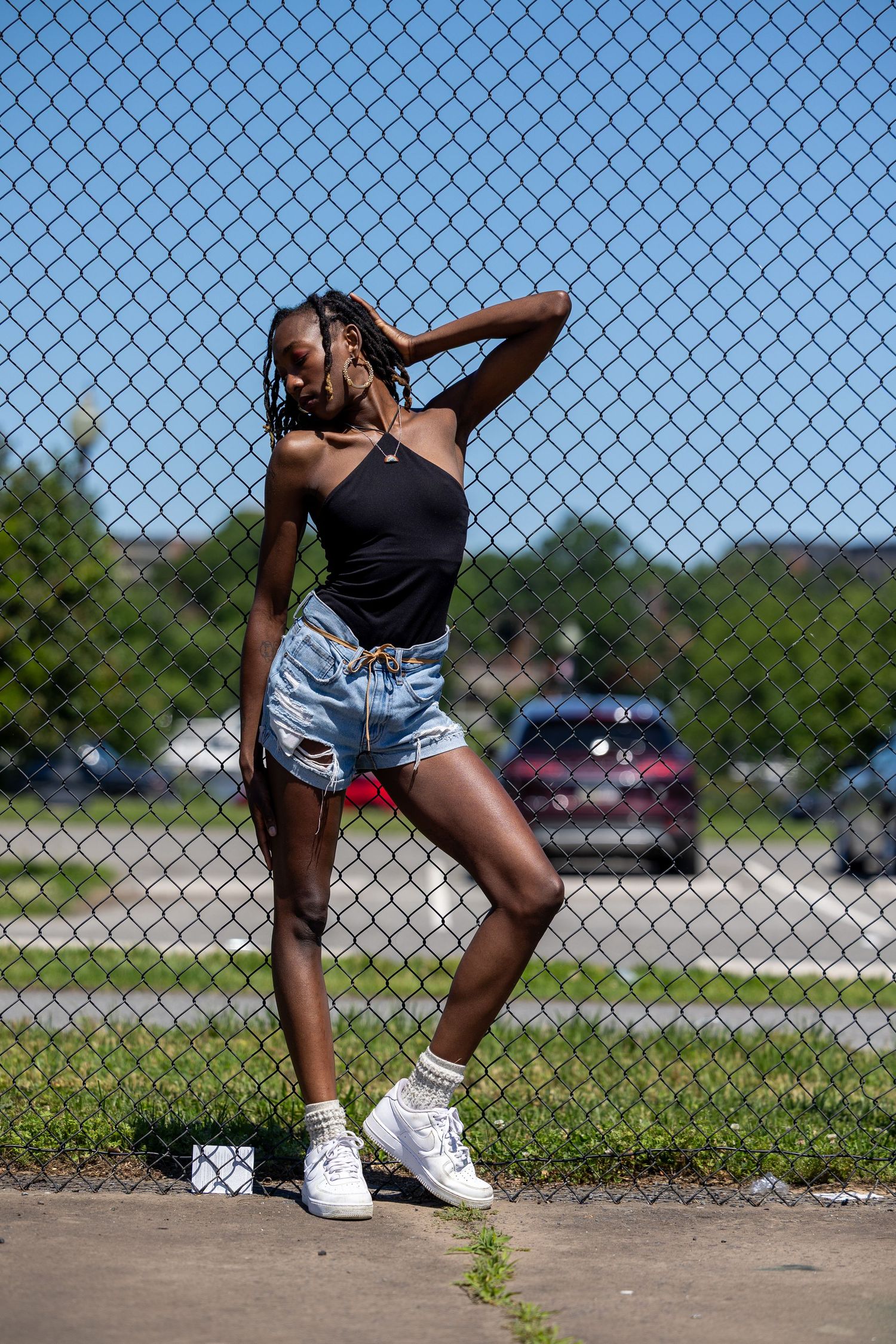 Young woman in black top and denim shorts posing against chain-link fence outdoors on sunny day.