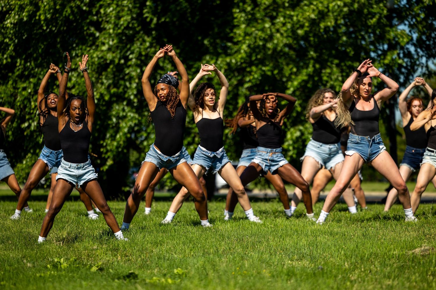 Group of young women in black tops and denim shorts performing a synchronized dance routine outdoors in a park.